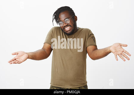 Piscina colpo di bella incurante afro-americano con la barba e taglio di capelli alla moda, sollevando le palme e scrollare con clueless gesto, essendo inconsapevoli e indifferente, in piedi felice su muro grigio Foto Stock