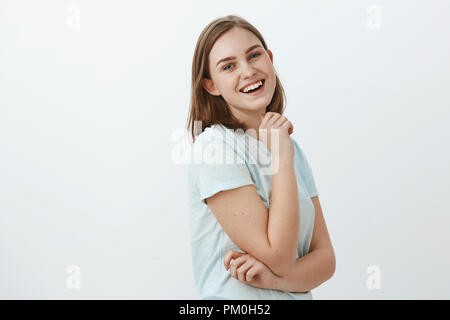 Studio shot di abili creativi e ambiziosi donna europea nel quartiere alla moda di t-shirt in piedi di profilo su muro bianco girando alla fotocamera con soddisfatto felici e sicuri di sé sorriso tenendo la mano sul mento Foto Stock