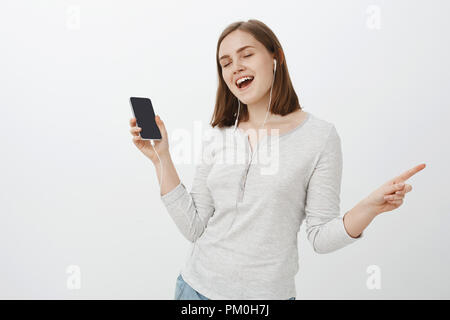 Gioiosa ed entusiastica di buona ricerca europea femminile marrone con taglio di capelli chiudendo gli occhi che porta lontano dal divertimento e gioia mentre si ascolta la musica in cuffia tenendo lo smartphone su muro grigio Foto Stock