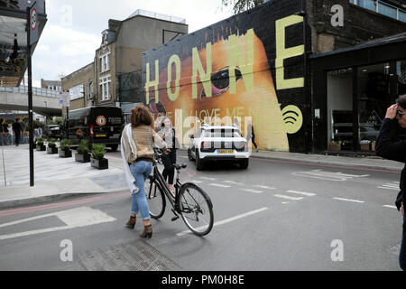 Donna che cammina con la bici in strada dal cittadino M Hotel passato HONNE carta murale street art pittura su Holywell Ln Shoreditch East London REGNO UNITO KATHY DEWITT Foto Stock