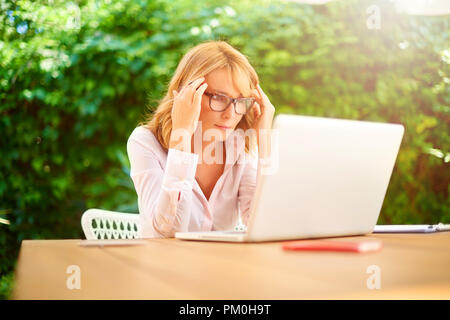 Colpo di donna di mezza età con un mal di testa mentre è seduto alla scrivania e di lavoro sul computer portatile. Home office. Foto Stock