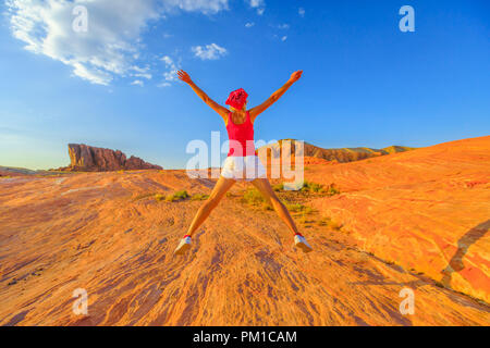 Giovane ragazza escursionismo jumping a valle del fuoco del parco statale, Nevada, Stati Uniti. Felice escursionista cercando il paesaggio a strisce verso l'alto la formazione al nord. Onda di fuoco sentiero al tramonto il colore rosso. Foto Stock