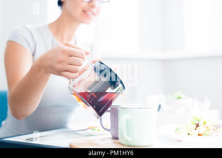 Primo piano di un irriconoscibile giovane donna versando il tè nero dal bollitore di vetro mentre vi gustate il pranzo nel soleggiato cafe, spazio di copia Foto Stock