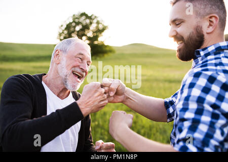 Un adulto hipster figlio col padre senior rendendo fist bump in natura al tramonto. Foto Stock