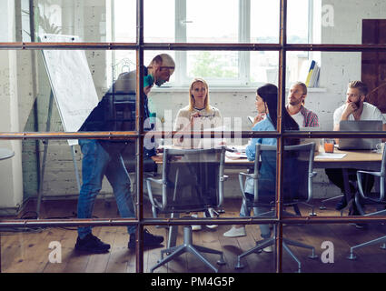 Giovani professionisti team. Un gruppo di giovani uomini moderni in smart casual avente una riunione di brainstorming in piedi dietro la parete di vetro nell'ufficio di creative. Foto Stock