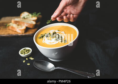 Crema di zucca zuppa nel vaso su sfondo nero. Decorazione a mano minestra con erbe aromatiche. Alimentare il concetto di styling Foto Stock