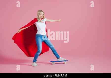 Il supereroe su uno skateboard. Funny giovane donna in immagine di un supereroe rides uno skateboard. Sfondo rosa Foto Stock