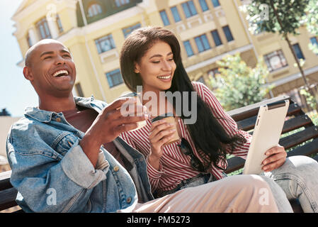 L'utilizzo dei gadget. Diversi giovani giovane seduto su un banco di lavoro sulla strada di città poggiarvi bicchieri di bere il caffè caldo donna navigare in internet sulla tavoletta digitale ridendo felice Foto Stock