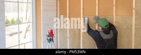 Il lavoratore fa opere di finitura delle pareti con un bianco tavola di legno, utilizzando una linea laser livello. Costruzione di isolante termico eco-telaio in legno casa con legno piastre in fibra Foto Stock