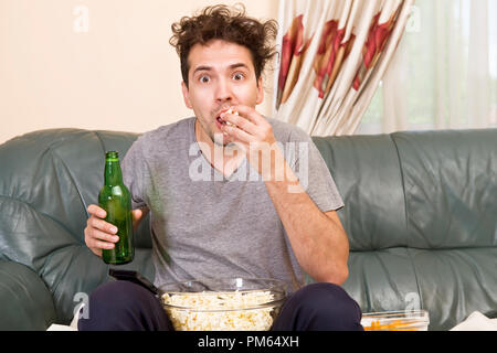 Uomo con birra e patatine guardando la TV a casa Foto Stock