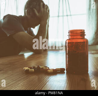 Uomo maturo che tiene testa con bottiglia di farmaco sul tavolo. Depressione maschile/salute mentale/malattia, PTSD... immagine concettuale. Foto Stock