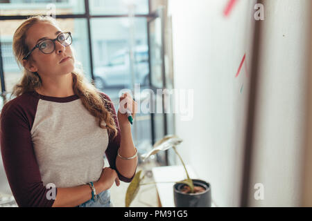 Giovane donna in piedi in ufficio e guardando la scheda di presentazione del pensiero. Femmina programmatore computer pensando su nuovo piano di progetto al tech start Foto Stock