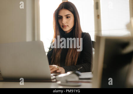 Ritratto di giovane e bella donna seduta alla sua scrivania in ufficio e di lavoro sul computer portatile. Smart giovane femmina in casuals lavorando sul computer portatile in ufficio Foto Stock
