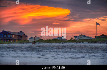 Giugno 18, 2018 Wrightsville Beach, NC: un surfista passeggiate lungo la sabbia sulla Wrightsville Beach come il sole tramonta in background. Foto Stock