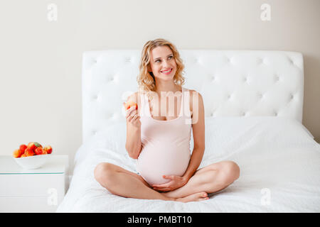 Ritratto di sorridere bianca bionda caucasica donna incinta azienda frutta dolce. Aspetta felice giovane età media femmina signora seduta sul letto in camera da letto Foto Stock