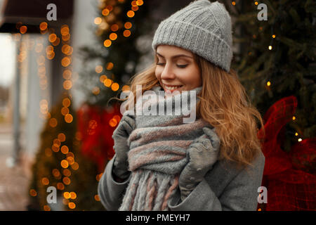Bella elegante giovane donna sorridente in maglia abiti alla moda con un cappello e una sciarpa su una vacanza vicino le luci e un albero di Natale sulla neve Foto Stock