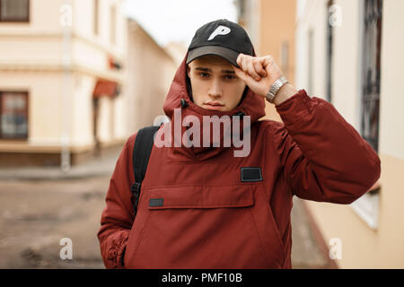 Moda giovane in un rosso giacca elegante con uno zaino regola un cappello da baseball sulla strada in città Foto Stock