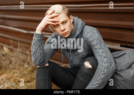 Elegante alla moda giovane uomo in una maglia maglione grigio con uno zaino seduto vicino a una parete arrugginito Foto Stock