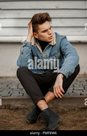 Bellissimo modello di moda un uomo con un taglio di capelli in una zona alla moda denim giacca con pantaloni neri e scarpe seduto vicino a una parete in legno Foto Stock