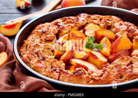 Comfort food dessert: peach sgretolarsi in un tondo nero piatto di cottura, vicino, con pesche fresche dietro il piatto Foto Stock