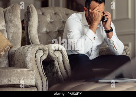 Ritratto di sottolineato imprenditore maturo seduto in una stanza di albergo e parlando al telefono. Imprenditore seduto sul divano con la mano sulla testa parlando sulla cella p Foto Stock