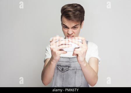 Arrabbiato crazy gamer adolescente che indossa in demin tuta e T-shirt bianca con piedi, utilizza lo smartphone e la riproduzione di gioco mobile con turbato viso. Indoor, iso Foto Stock