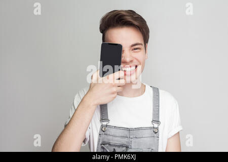 Ritratto di divertenti moderno bello giovane ragazzo in demin tuta e T-shirt bianca con piedi, di tenuta e di copertura di un occhio con lo smartphone, guardando a camma Foto Stock