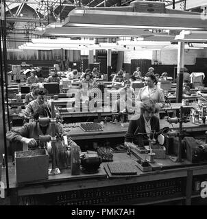 Anni '1959, lavoratrici storiche sedute su lunghe panche di legno, utilizzando piccole macchine industriali, nell'officina della fabbrica di batterie Ever Ready di Walthamstow, Londra, Inghilterra, Regno Unito. Nella foto è visibile una supervisore donna che controlla il lavoro dei dipendenti. Foto Stock