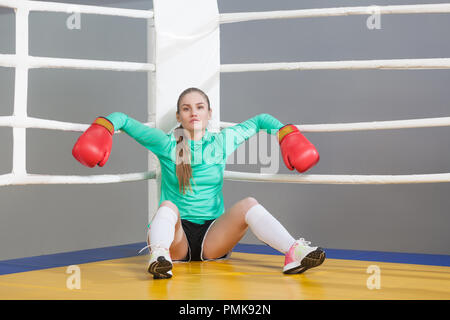 Ritratto di fiducioso bella giovane atleta donna con i capelli raccolti di posa e appoggiata in un angolo del pugilato ring in rosso guanti e guardando a c Foto Stock