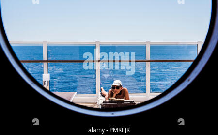 La donna a prendere il sole e la lettura sul ponte, visto attraverso una nave tonda finestra sulla nave da crociera MSC Seaview Foto Stock
