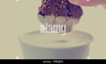 Persona dunking un piccolo biscotto croccante in una tazza di latte schiumoso o cappuccino con schiuma aderente al cookie in un retrò effetto tonico. Foto Stock