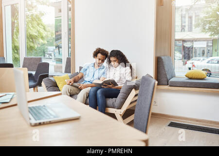 Coppia di bella arte multietnica gli studenti in abiti casual seduto comodo divano in grande e luminosa libreria, la lettura di informazioni sulla storia di film in libri, ottenere pronto per l'esame. Foto Stock