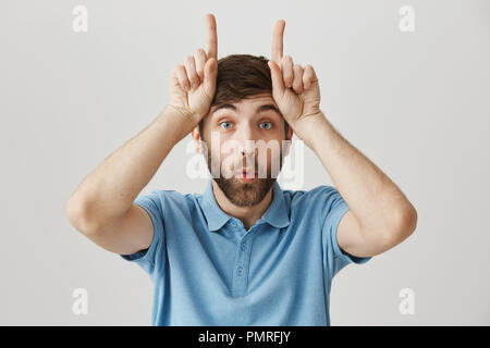 Close-up verticale di positivo divertenti giovane ragazzo con barba, rendendo volti e tenendo le dita indice oltre il fronte rivolto verso l'alto o per la visualizzazione di capra o le corna, dicendo moo e piedi su sfondo grigio Foto Stock