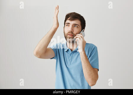 Guy non può richiamare qualche momento. Studio shot di perplesso e pensieroso giovane ragazzo parlando sullo smartphone mentre guardando in alto e gesticolando con aumentata palm, cercando di ricordare il nome, in piedi su muro grigio Foto Stock