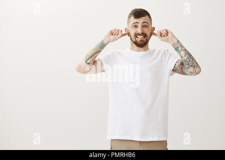 Che cosa terribile rumore. Studio shot di orinato scomodo bello uomo barbuto con tatuaggi, coprendo le orecchie con le dita indice e smorfie da antipatia, udito orribile riproduzione di un amico Foto Stock