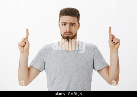Ritratto di scontento triste e sciocco bello maschio con la barba e baffi accigliata rendendo tetro sorriso rivolto verso l'alto esprimendo il proprio rammarico o gelosia indignazione permanente contro lo sfondo grigio Foto Stock