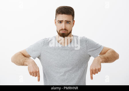 Vita-up shot di cupo maschile carino il ragazzo con la barba e capelli castani accigliata e busbana francese con sciocche infelice la faccia rivolta verso il basso sentirsi soli e triste dopo ingiusto cosa è accaduto oltre il muro grigio Foto Stock