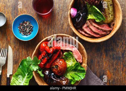 Bistecca alla griglia con verdure alla griglia servita nella piastra di pane sul tavolo di legno con un bicchiere di vino rosso, vista dall'alto Foto Stock