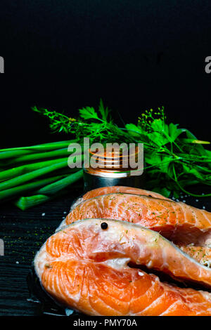 Fette di pesce rosso sul salmone trota con fresco verde cipolla, prezzemolo, peppper, spezie, aneto, rosa e bianco sale, selery verdi su nero backgrou in legno Foto Stock