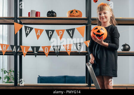 Adorable kid con la zucca in piedi sulla scaletta a appendere bandiere con happy Halloween iscrizione a casa Foto Stock