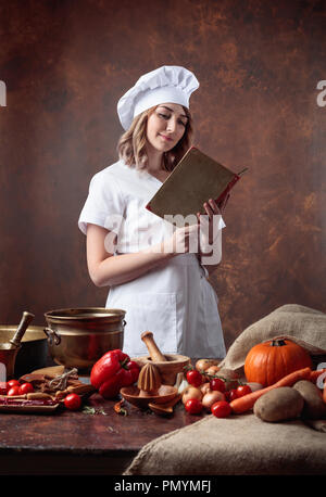 Giovane bella ragazza in un chef uniformi della legge un vecchio libro di cucina. Sul tavolo, diversi utensili da cucina e verdure. Foto Stock