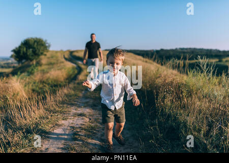 Adorabili happy little boy in esecuzione sul percorso rurale mentre il padre a piedi dietro Foto Stock