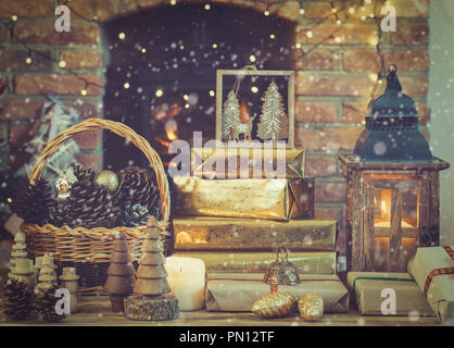 Tonico composizione di Natale con creato neve, lanterna, presenta decorazioni sul tavolo davanti al camino con woodburner, ornamenti di legno e garl Foto Stock