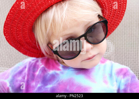 Un piccolo grazioso 2 anno vecchia ragazza toddler è prendere il sole al di fuori, indossando un rosso estate cappello e occhiali da sole scuri, come ella loos la fotocamera a effettuare un serio Foto Stock