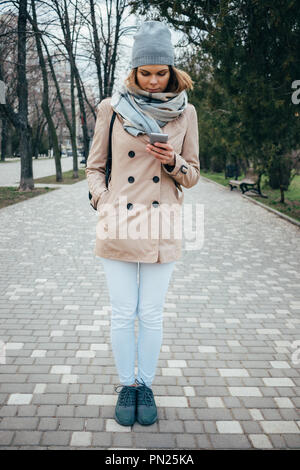 Full lenght verticale Ritratto di giovane donna che indossa il beige trench coat, sciarpa e cappello utilizzando smart phone nel parco della città. Donna che guarda la sua mobil Foto Stock