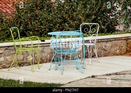 Un tavolo e sedie nel giardino. Acciaio colorato un tavolo e le sedie in piedi in un bellissimo giardino alla giornata di sole. Mobili da giardino. Un set di tre colorati. Foto Stock