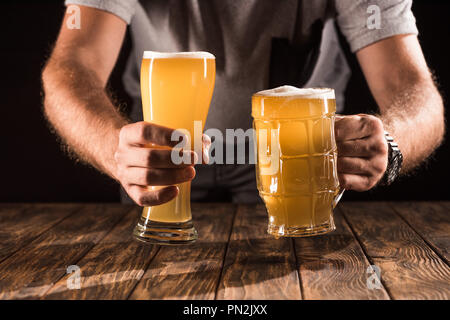 Immagine ritagliata dell uomo di mettere due tazze con birra fresca con schiuma al tavolo di legno Foto Stock
