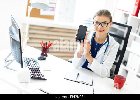 Una giovane ragazza in un abito bianco si siede a un tavolo in ufficio ed è in possesso di un telefono cellulare in mano. Uno stetoscopio appeso attorno al collo. Foto Stock