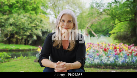 Il vecchio felice white donna seduta nel lussureggiante giardino caldamente guarda la fotocamera Foto Stock