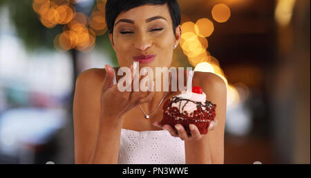 Sorridente femmina nero mangiare cupcake fantasia in ambiente esterno con luci bokeh di fondo Foto Stock